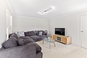 A modern living room with a sectional sofa, a glass coffee table, a flat-screen TV on a wooden cabinet, and a ceiling light fixture.