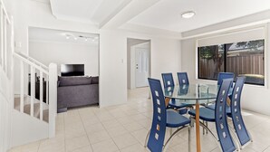 A modern kitchen with white cabinetry, a dining area with a glass table and blue chairs, and a staircase leading to another level. - Fingal Sands 10A - Fingal Bay (Fingal Bay)
