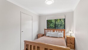 A bedroom with a wooden bed, a nightstand with a lamp, a window with a view of trees, and a white door. - Fingal Sands 10A - Fingal Bay (Fingal Bay)