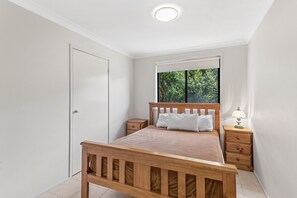 Een slaapkamer met een houten bed, een nachtkastje met een lamp, een raam met uitzicht op bomen, en een witte deur.
