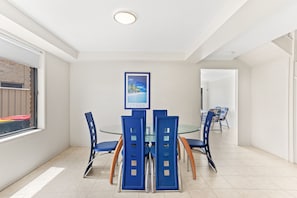 A dining area with a glass table surrounded by blue chairs, a framed picture on the wall, and a view of the outside through a window.