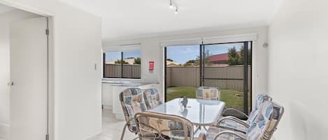 A dining area with a table and chairs, a kitchen in the background, and a view of the backyard through glass doors.