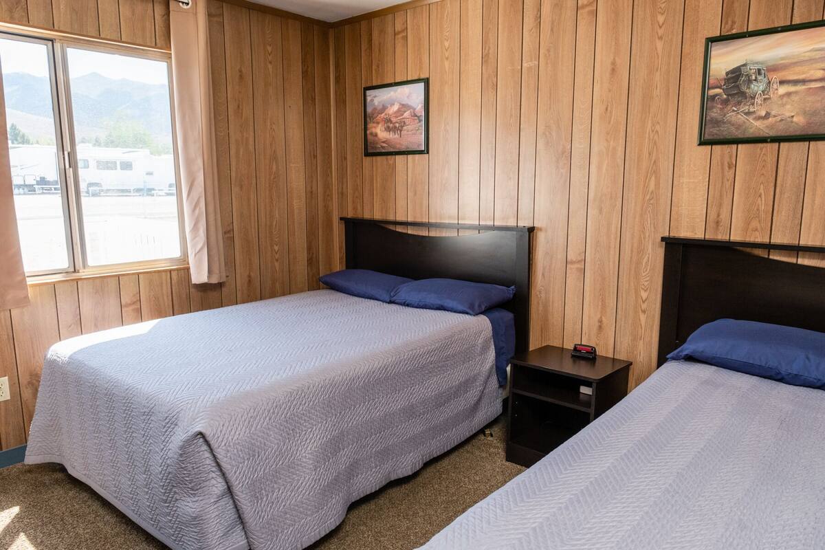A room with wooden panel walls, two beds with blue pillows, a nightstand, and a painting on the wall.