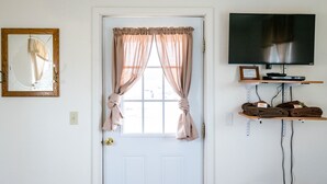 Quarto com porta branca, cortinas bege amarradas, espelho emoldurado, televisão de tela plana e prateleira com toalhas.