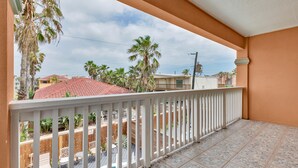 Ein Balkon mit weißer Reling, gefliestem Boden und Blick auf Palmen und Häuser im Hintergrund.