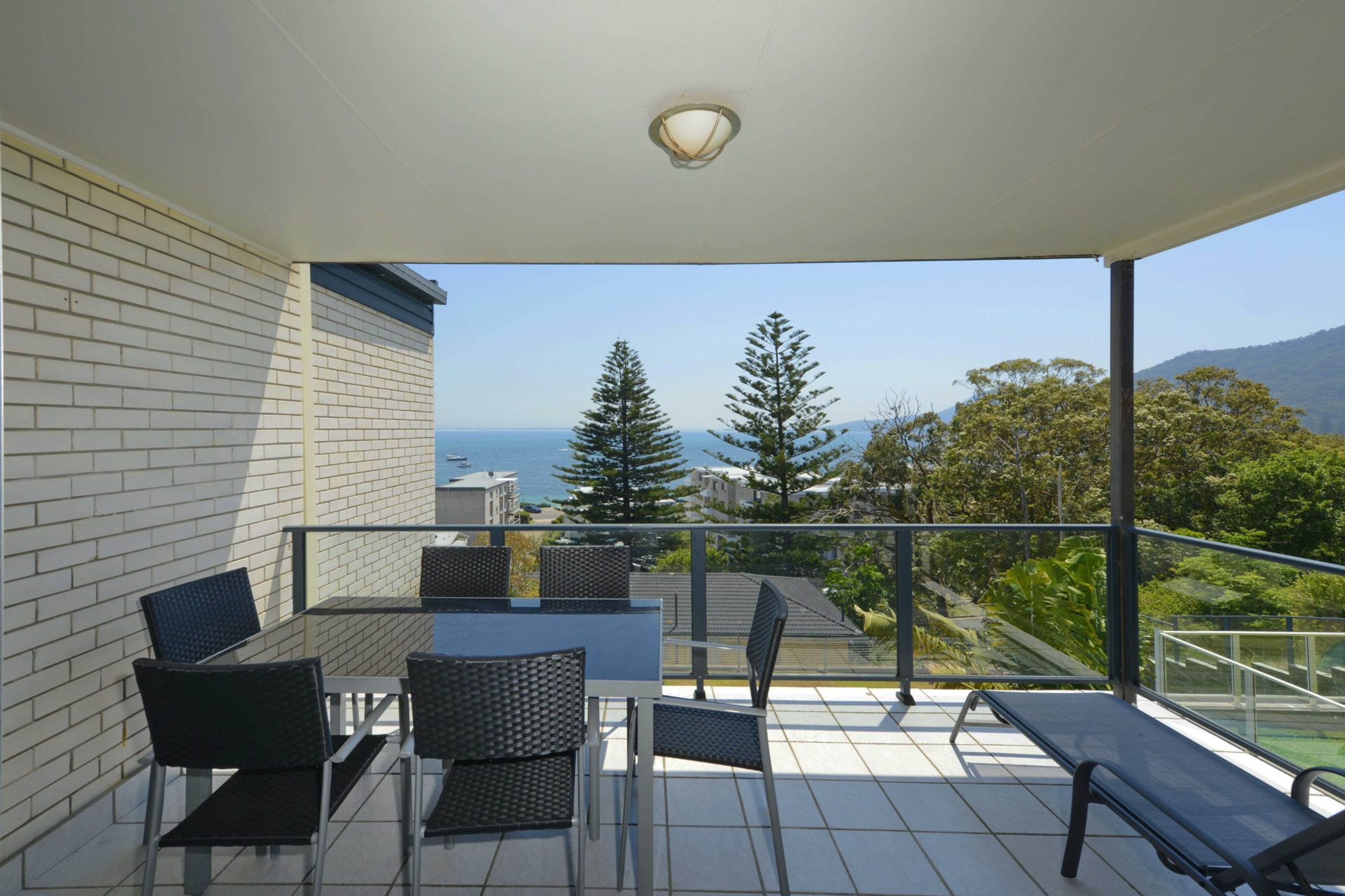 A balcony with a table and chairs, a bench, and a view of the sea and trees.
