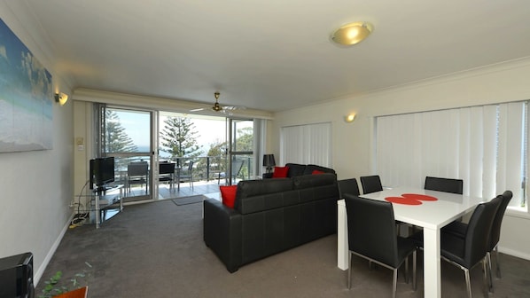 Una sala de estar moderna con un sofá negro, una mesa de comedor con sillas y un balcón con vistas a árboles y al mar.