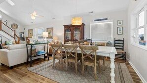 A spacious living room with a dining area, wooden flooring, a white ceiling fan, and a large window with blinds. - Oceanfront Heaven's Anchor in Holden Beach, 3BR, 2BA, perfect for family fun! (Holden Beach)