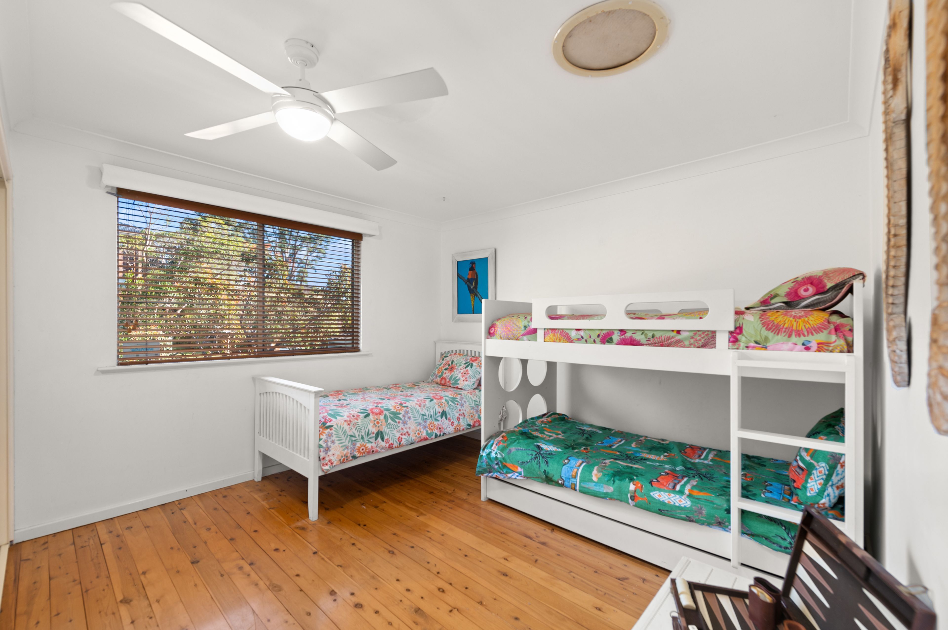 A room with a bunk bed, a single bed, a ceiling fan, a window with blinds, and a small white table.