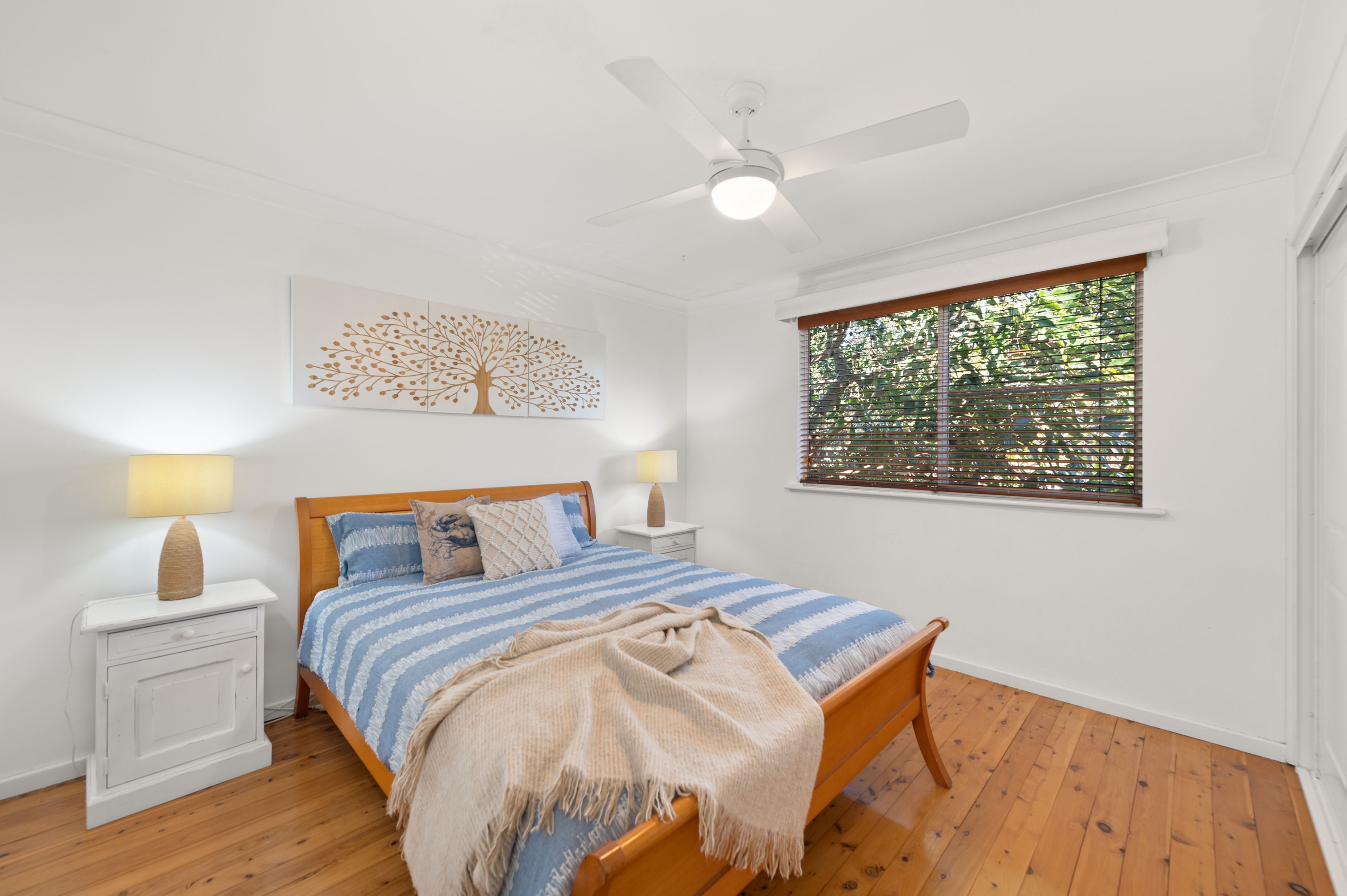 A bedroom with a bed, two bedside tables, a ceiling fan, a window with blinds, and a wall-mounted artwork.