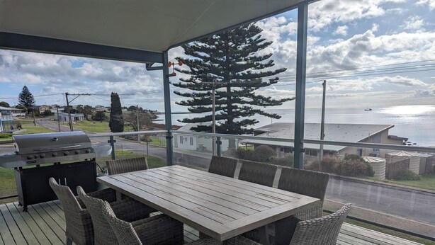 A covered outdoor area with a table and wicker chairs, overlooking a body of water and a road. - Woody's Beach House ~ Beautiful Sea Views, Great Position,  Off Peak Special! (Point Turton)