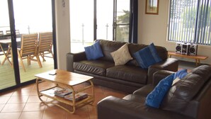 A living room with a brown leather sofa, a wooden coffee table, and a television. There is a window with blinds and a view of the outside. - Woody's Beach House ~ Beautiful Sea Views, Great Position,  Off Peak Special! (Point Turton)