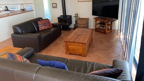 A living room with a sofa, a coffee table, a TV, a fireplace, and a kitchen area in the background. - Woody's Beach House ~ Beautiful Sea Views, Great Position,  Off Peak Special! (Point Turton)