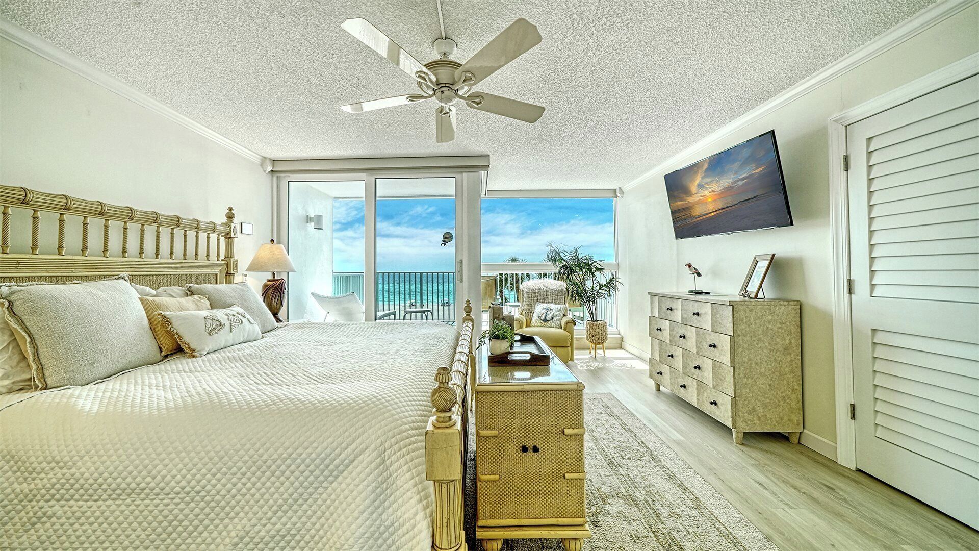 A bedroom with a large bed, a ceiling fan, a view of the ocean, and a television on the wall.