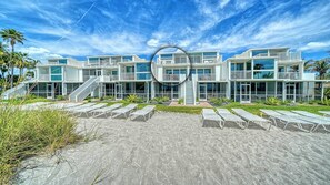 Una moderna propiedad frente a la playa con varios balcones, una escalera central y una zona de arena con tumbonas.