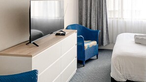 A hotel room with a flat-screen TV on a wooden desk, a blue upholstered chair, a white bed, and a grey carpet.