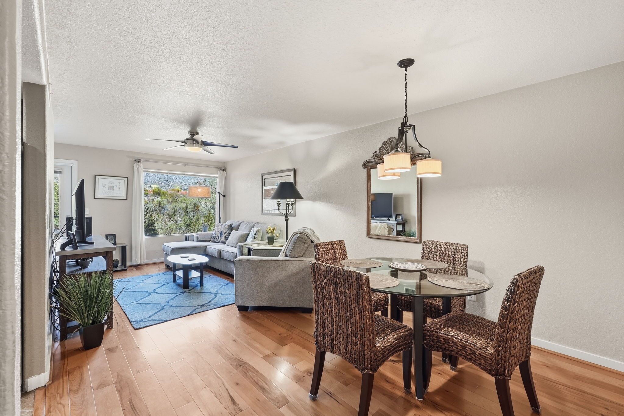 A modern living room with a dining area, a ceiling fan, and a large window offering a view of greenery.