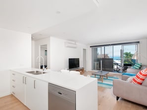 A modern kitchen with a dishwasher, a dining area with a table and chairs, a sofa with striped pillows, and a large sliding glass door leading to a balcony with a view. - The Shoal 210 Modern Apartment in Shoal Bay (Shoal Bay)