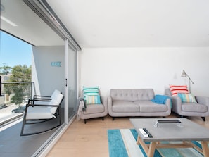 A balcony with a sofa, armchair, and a small table with a remote control. - The Shoal 210 Modern Apartment in Shoal Bay (Shoal Bay)