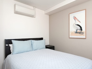 A bedroom with a bed, pillows, a nightstand, a wall-mounted air conditioner, and a framed painting of a pelican. - The Shoal 210 Modern Apartment in Shoal Bay (Shoal Bay)