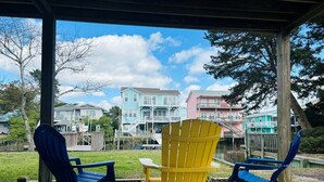 A covered outdoor seating area with Adirondack chairs and a wooden table, overlooking a residential area with colorful houses. - Dry Dock / 150 Tarpon: Spacious 4-bedroom home in Holden Beach with canal views. (Holden Beach)