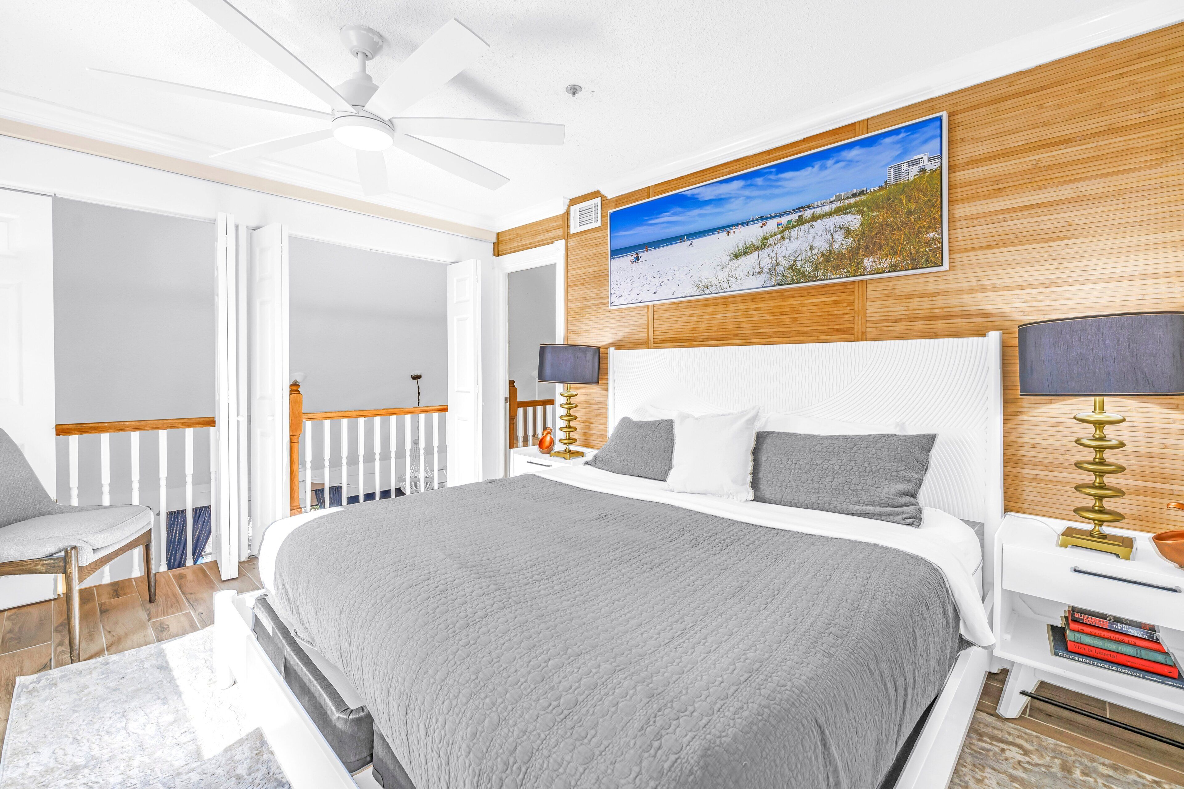 A bedroom with a large bed, a ceiling fan, a wooden headboard, a TV, bedside lamps, and a beach-themed picture on the wall.