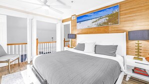 Ein Schlafzimmer mit einem großen Bett, einem Deckenventilator, einem Holz-Kopfteil, einem Fernseher, Nachttischlampen und einem Bild mit Strandmotiv an der Wand.