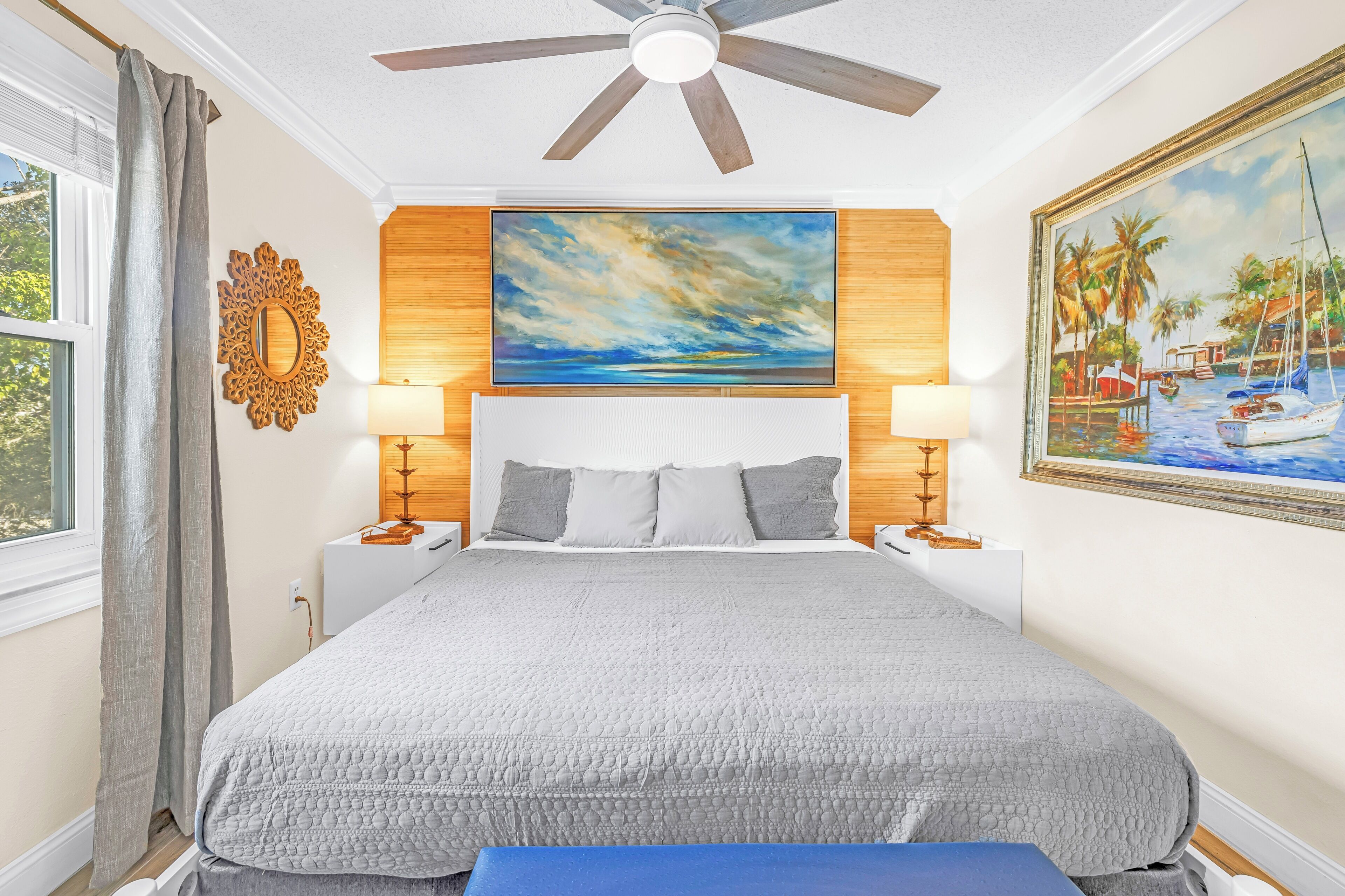 A bedroom with a large bed, a ceiling fan, two bedside lamps, a painting, and a window with curtains.