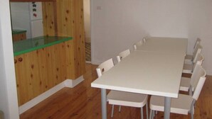Ein Zimmer mit einem langen Tisch, weißen Stühlen, Holzvertäfelung an einer Wand und einer Küchenzeile.