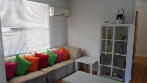 Ein Wohnzimmer mit einem beigefarbenen Sofa, bunten Kissen, einem weißen Couchtisch, einem weißen Beistelltisch und einer weißen Regalwand mit Dekorationselementen.