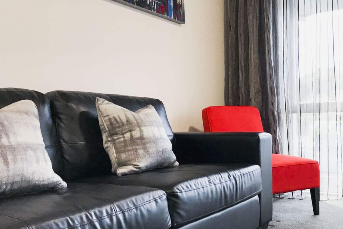 A black leather sofa with two pillows and a red chair in a room with a large window and curtains.