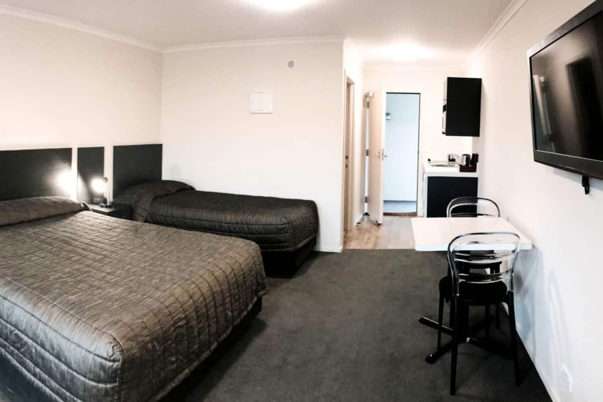 A hotel room with two beds, a desk, a chair, a TV, and a small kitchenette.