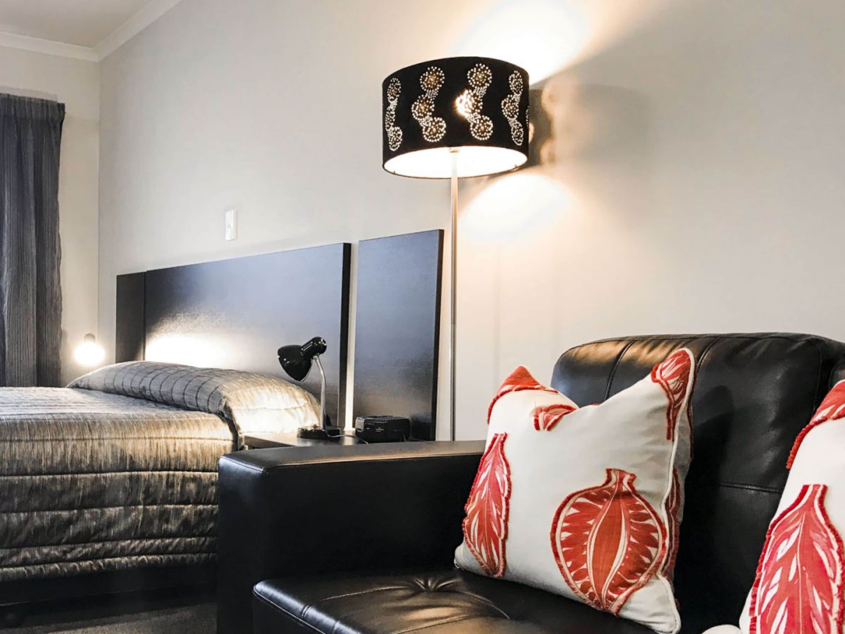 Habitación de hotel con cama, sofá de cuero negro y cojines estampados en rojo y blanco, lámpara negra con pantalla decorativa y cabecera moderna.