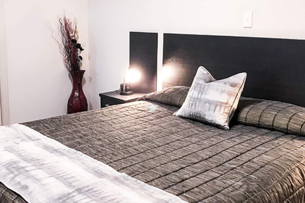 A neatly made bed with a quilted headboard, a bedside table with a lamp, and a vase with dried branches on the nightstand.
