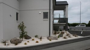 A modern building with a flat facade, a small garden area with rocks and plants, and a parking lot with a white car.