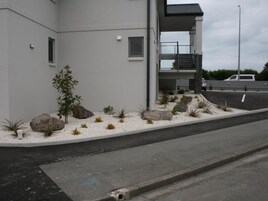 Un edificio moderno con fachada plana, un pequeño jardín con rocas y plantas, y un estacionamiento con un auto blanco.