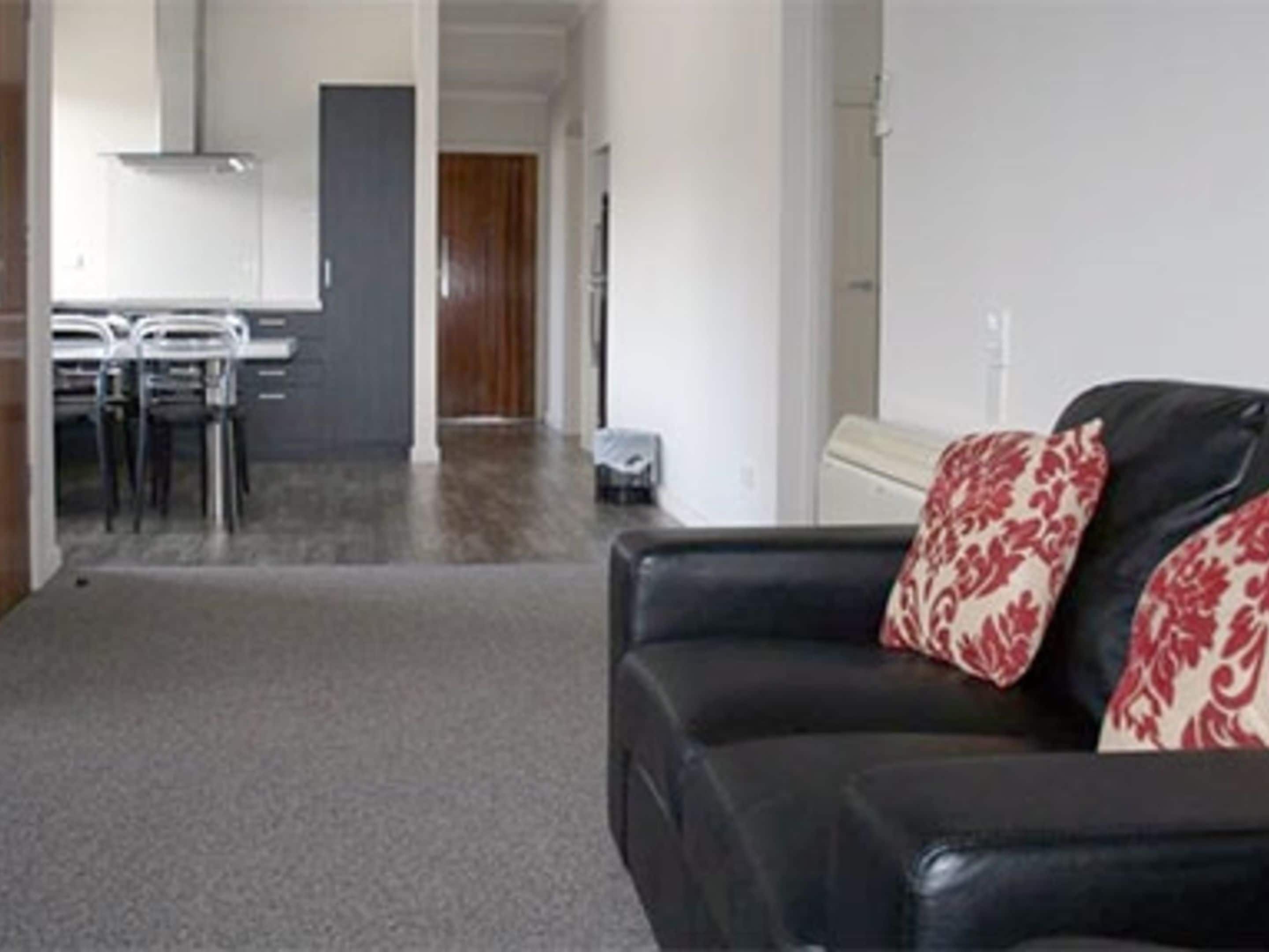 Una sala de estar con un sofá de cuero negro, una zona de comedor con sillas y una mesa, y una cocina al fondo.