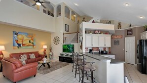 A spacious living room with a red leather sofa, a coffee table, and a TV. There is a kitchen area with white cabinets, a refrigerator, and a bar counter. - 6BR Pool Home Windsor Palms by SHV-2238 (Kissimmee)