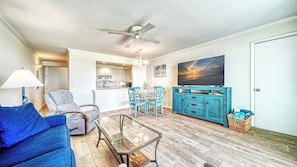 Un salon avec un canapé, une table basse en verre, un coin repas avec des chaises, une cuisine, un ventilateur de plafond et un tableau accroché au mur.