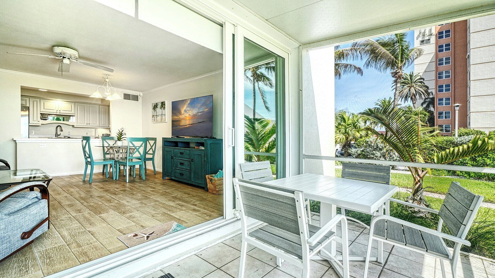 A modern kitchen with white cabinets, a dining area with teal chairs, a TV on a teal cabinet, and a balcony with a view of palm trees and buildings.