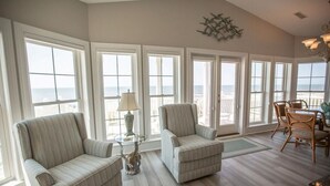 A spacious room with large windows offering a view of the ocean, two upholstered chairs, a small table with a lamp, and a dining area with chairs. - Paw-Paw's Place: Spacious 6BR oceanfront home in Holden Beach, sleeps 14! (Holden Beach)