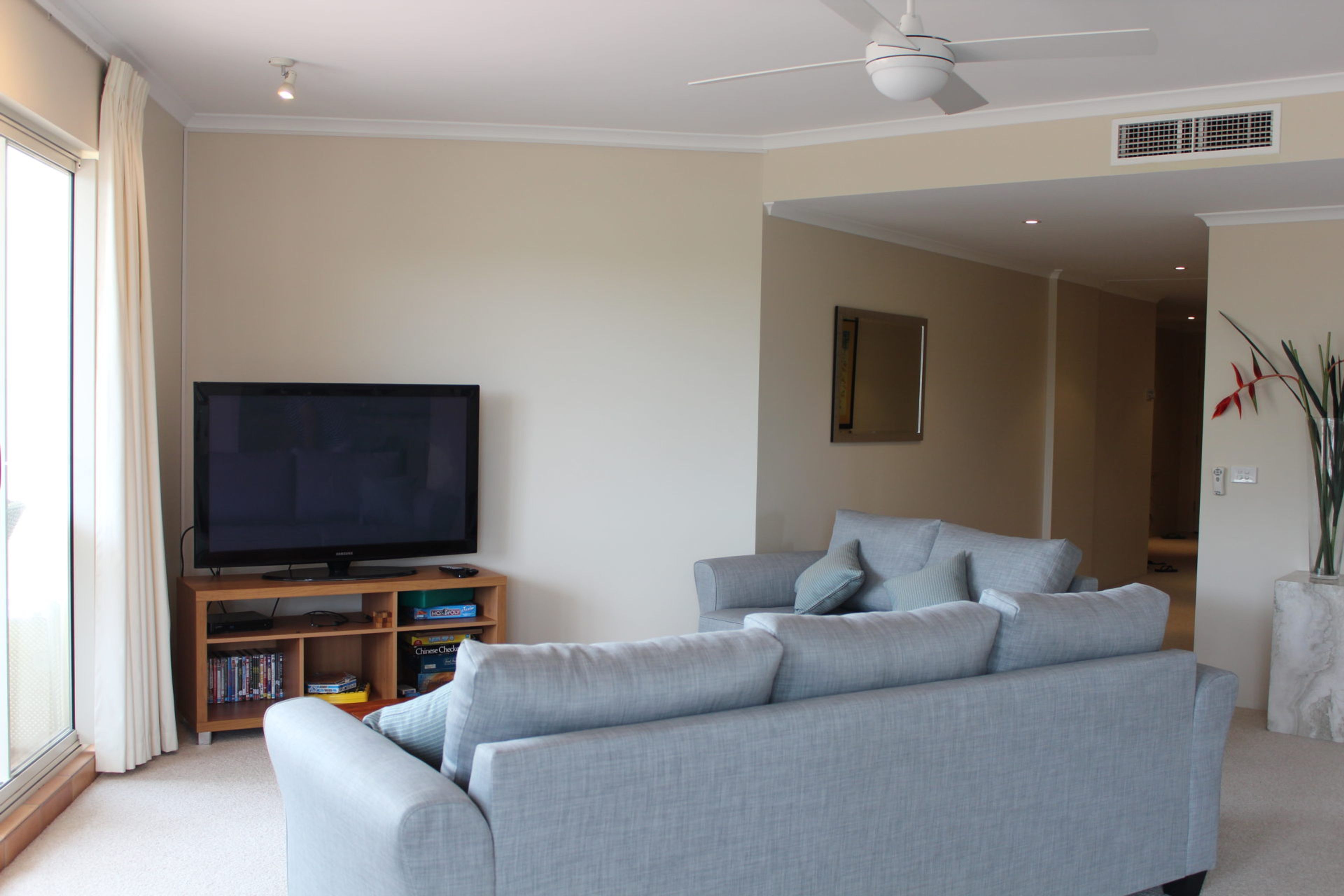 A living room with a flat-screen TV, a grey sofa, a bookshelf, and a ceiling fan.