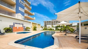 Área de piscina com céu azul e um edifício de vários andares com varandas, e mobiliário de exterior com guarda-sóis.