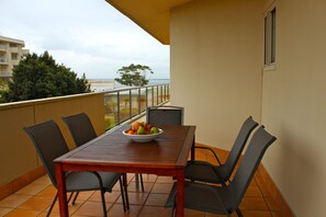 A balcony with a table and chairs, a bowl of fruit, and a view of the sea and buildings.
