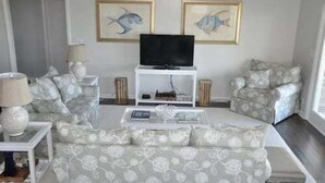 Uma sala de estar com sofá, poltronas, mesa de centro, televisão em móvel e dois quadros de peixes emoldurados na parede.