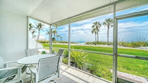 A covered outdoor area with a table and chairs, overlooking a beach with palm trees and a clear sky.
