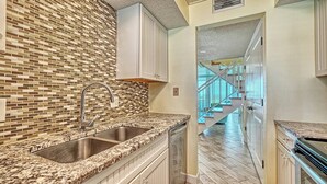 Eine Küche mit gefliestem Rückspritzschutz, Granit-Arbeitsplatten, einer Edelstahl-Spüle und Blick auf eine Holztreppe, die zu einem Balkon führt.