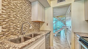 Eine Küche mit gefliestem Rückspritzschutz, Granit-Arbeitsplatten, einer Edelstahl-Spüle und Blick auf eine Holztreppe, die zu einem Balkon führt.