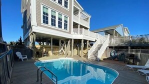 Una terraza de madera con piscina, mobiliario exterior y un edificio de varios niveles con balcones.