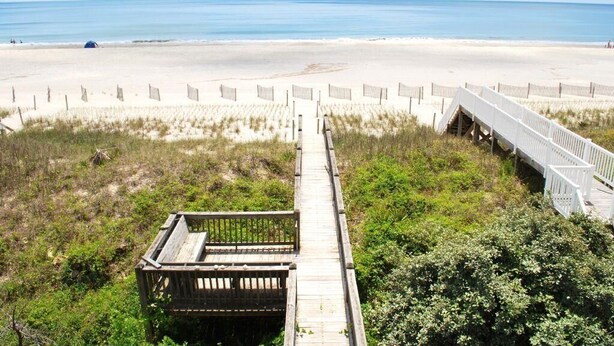 A beach with a wooden boardwalk leading to the shore, surrounded by sand and vegetation. - Oceanfront bliss awaits at Our Sand Dune, a spacious 5-bedroom retreat in Holden Beach. (Holden Beach)
